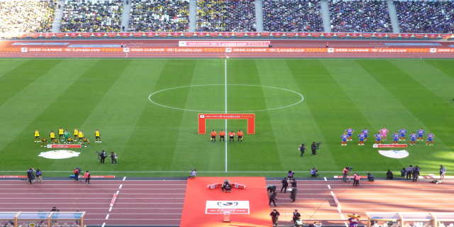 Eine Gruppe von Menschen steht auf einem grünen Fußballfeld, umgeben von einem Stadion voller Zuschauer, mit Werbetafeln, die wahrscheinlich für das 2020 Levin Cup-Finale werben.
