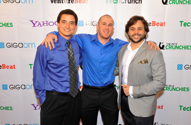 Drei Personen vor einem Sponsorenbanner, alle mit Kleidung.