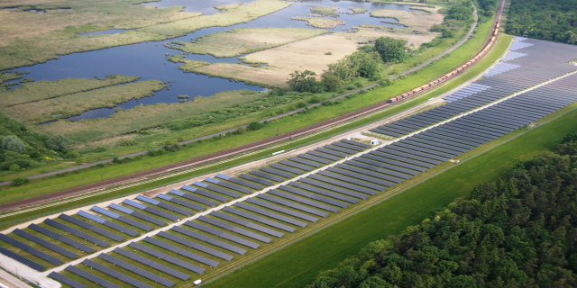 Luftaufnahme einer Solar-Farm mit Panelen, umgeben von Bäumen, Gras, Wasser und einer Bahn auf einem Gleis.