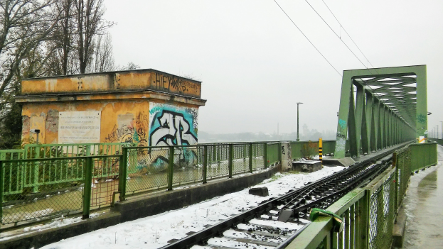 Eisenbahnschiene mit Graffiti an der Seite, umgeben von Geländern, Pfählen, Lampen, Drähten und Bäumen, mit Schnee bedeckt und sichtbarem Himmel im Hintergrund.