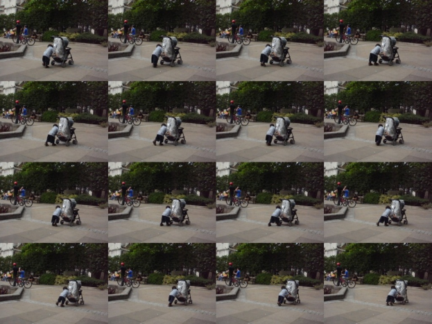 Ein Kind geht auf einer Straße und zieht einen Karren, mit zwei Personen, die links Fahrräder halten, Bäumen im Hintergrund und einem Gebäude links.