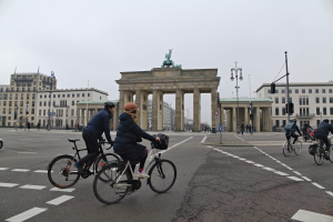 Eine Gruppe von Radfahrern in verschiedenenfarbiger Kleidung und Helmen fährt eine Straße vor dem Brandenburger Tor in Berlin, Deutschland, entlang, mit Laternen, Ampeln und einer Menschenmenge im Hintergrund.