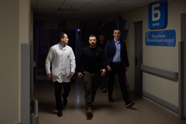 Eine Gruppe von Männern geht einen Krankenhausflur entlang, sieht besorgt aus, mit einer Tür auf der rechten Seite und Texttafeln auf der linken Seite.