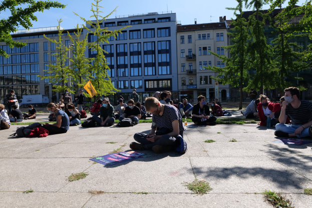 Eine Gruppe von Menschen sitzt vor einem Gebäude auf dem Boden während einer Demonstration in Berlin, einige tragen Masken und sind von Taschen und Bäumen umgeben unter einem klaren blauen Himmel.