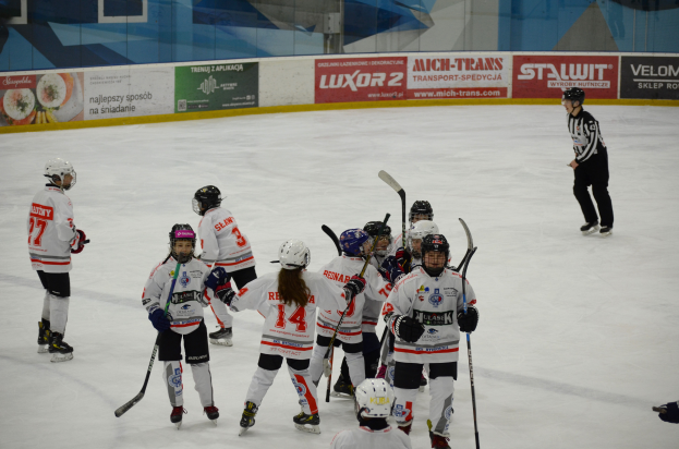 Eine Gruppe junger Menschen, die auf einem Eisplatz Eishockey spielen, alle tragen Helme und halten Hockeystöcke, mit einer Person, die rechts steht und Brettern mit Text im Hintergrund.