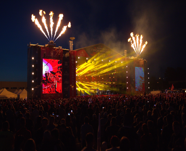 Eine große Menschenmenge bei einem Musikfestival schaut auf einer hell erleuchteten Bühne mit Bannern, umgeben von Zelten und Feuerwerk am dunklen Himmel.