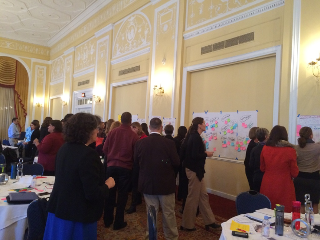 Eine Gruppe von Menschen untersucht Plakate und Charts, die an einer Wand in einem Gebäude zu sein scheinen, das wie ein Kunstmuseum aussieht.