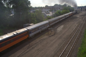 Ein Zug auf Schienen mit Bäumen, Strommasten mit Kabeln, Autos und Gebäuden links, eine Brücke davor und Rauch aus dem Zug.