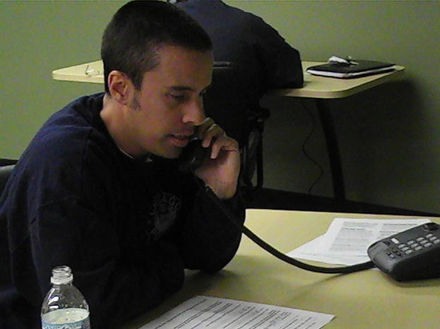 Mehrere Personen sitzen um einen Tisch, wobei eine Person mit einem auf dem Tisch liegenden Telefon spricht, neben einer Station, einer Einwegflasche und Kabeln.