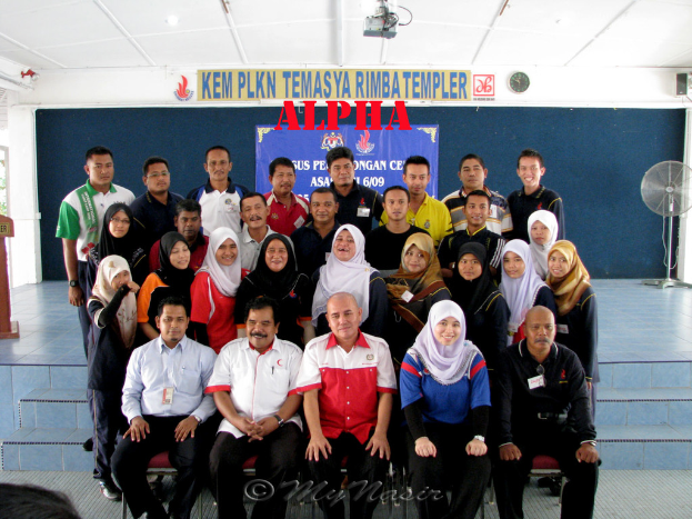 Eine Menschenmenge, die lachend vor einer Bühne posiert, mit einer Wandtafel im Hintergrund, einem Tischventilator rechts, einem Podium links und einem Projektor oben im Bild.