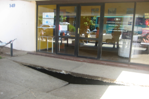 Eine gebrochene Oberfläche im Vordergrund mit einer Glastür, Wand und Pfahl im Hintergrund; die Spiegelung in der Glasscheibe zeigt Fahrzeuge auf der Straße und einige stehende Personen.