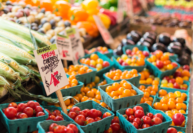 Ein Bauernmarkt mit Körben voller frischer Produkte wie Tomaten und Mais sowie Tafeln mit Text im Hintergrund.