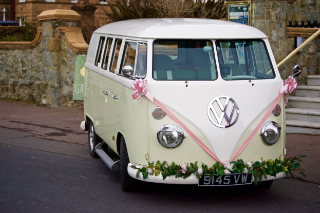 Ein weißer und rosafarbener VW-Bus mit floralen Verzierungen, der vor einer Steinmauer geparkt ist, mit einer Treppe auf der rechten Seite und einem Gebäude im Hintergrund.