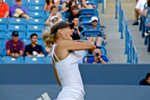 Eine Frau in einem weißen Oberteil und schwarzer Mütze steht vor einem blauen Stadionplatz mit Zuschauern.