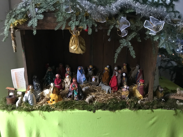 Eine Krippe mit einem Baby Jesus in einer Krippe umgeben von Spielzeugen, Gras und anderen Gegenst√§nden auf einem mit gr√ľner Decke bedeckten Tisch, mit einem geschm√ľckten Weihnachtsbaum im Hintergrund.