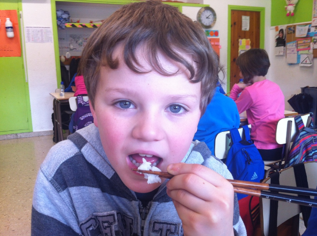 Ein Junge in einer grauen und schwarzen Jacke isst mit Stäbchen in einem Klassenzimmer, mit anderen Kindern im Hintergrund, einer grün-weißen Wand, Lehrplakaten und -tafeln an einer weißen Wand, einer braunen Tür und einer Wanduhr.