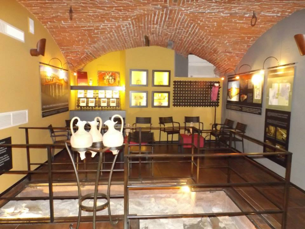 Ein Museumsraum voller Tische, Stühle, einem Geländer, Vasen, Texttafeln, Wandrahmen, Lampen und diversen Ausstellungsgegenständen.