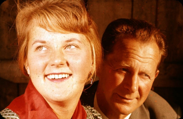 Ein Mann und eine Frau in inniger Umarmung, wobei der Mann seinen Arm um die Taille der Frau legt und ihr Kopf auf seiner Schulter ruht, vor einer schmucklosen Wand.