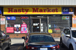 Ein geschäftiges Hasty Market-Geschäft in Toronto, Ontario, mit Autos auf der Straße davor; das Geschäft hat Glaswände, Türen und Plakate an den Innenwänden.
