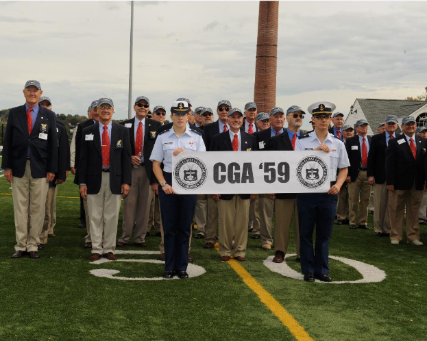Eine Gruppe von Männern in formeller Kleidung und Marineuniformen steht vor Häusern, mit Bäumen, Pfählen und einem bewölkten Himmel im Hintergrund.