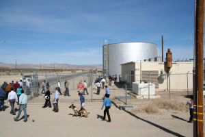 Eine spärlich besiedelte Landschaft mit wenigen Menschen, einem Hund, Häusern, Strommasten und einem Zaun unter einem klaren Himmel.
