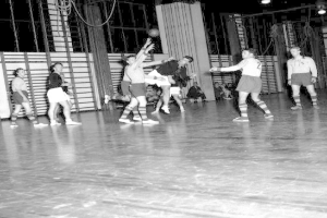 Eine Gruppe von Männern, die Handball auf einem Holzboden spielen, mit einer Wand und Lampen im Hintergrund, in Schwarz-Weiß.
