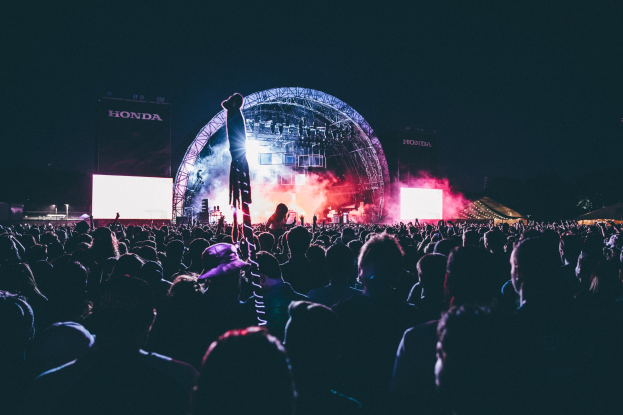 Eine große Menge Menschen auf einem Musikfestival mit einem Künstler auf der Bühne, umgeben von Musikern, beleuchtet von Lichtern, Bildschirmen und Bannern vor einem dunklen Hintergrund.