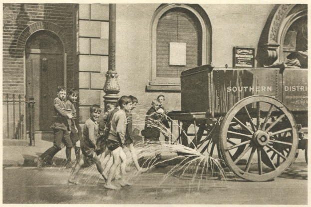 Schwarz-weiß-Bild von Kindern, die mit einem Hydranten vor einem Gebäude mit Fenstern, einer Tür, einem Mast und einem Zaun spielen, mit einem Wagen im Hintergrund.