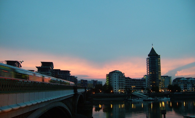 Ein Fluss mit weißen Booten, ein Zug auf einer Brücke rechts, Gebäude, Bäume, Lichter und Wolken im Hintergrund.