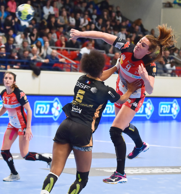 Eine Gruppe von Frauen, die auf einem Platz Handball spielen, während Zuschauer von Stühlen, Geländern und einer Wand im Hintergrund zuschauen.