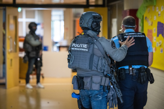 Zwei Polizisten stehen Seite an Seite in einem Flur, einer mit Helm und eine Waffe haltend, der andere am Boden, mit Papieren an der rechten Wand und Türen, Fenstern und anderen Gegenständen im Hintergrund.
