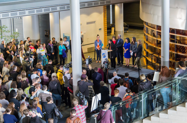 Gruppe von Menschen auf einer Treppe, die zu einem Podium mit Mikrofon führt, vor einer Menge mit Kameras, bei einem Event mit Schildern auf Ständern, Säulen, einer Tür und einem Topf mit einer Pflanze.