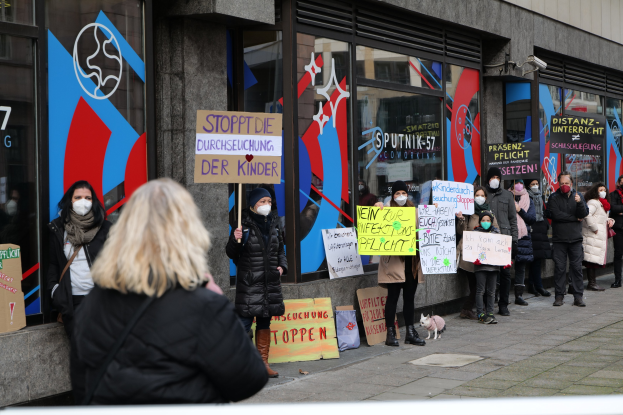Eine Gruppe von Menschen in Masken, die Schilder und Plakate halten, protestiert vor einem Gebäude mit Glaswänden und -türen, wobei Kameras und ein Hund unten zu sehen sind.