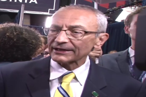 Ein Mann im Anzug mit Brille spricht zu Reportern über ein Mikrofonständer, umgeben von einer Menge, mit beleuchtetem Hintergrund und einer Tafel.