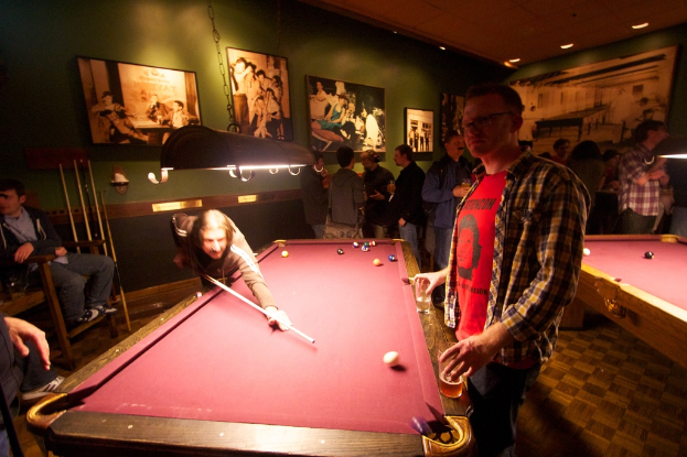 Zwei Personen spielen Snooker an einem von zwei Tischen in einem Club, mit mehreren Personen, die im Hintergrund stehen und sprechen, vor einer cremefarbenen Wand, die mit ein paar Fotorahmen geschmückt ist.