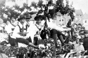 Eine Gruppe von Menschen in traditioneller bayrischer Tracht fährt auf einem mit Blättern geschmückten Motivwagen in einem Umzug mit, einige spielen Musikinstrumente, im Hintergrund sind Gebäude und ein klarer Himmel zu sehen, dargestellt in Schwarz-Weiß.