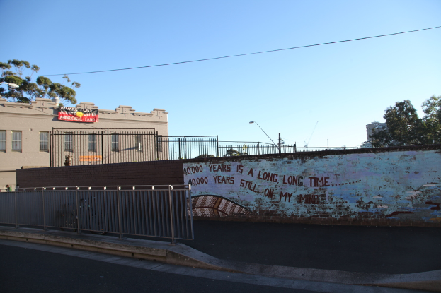 Eine Straßenszene in einer Stadt mit einer Straße, einem Geländer, Pflanzen, einer Wand, Text und einem Gehweg im Vordergrund, Bäumen, Gebäuden, Eisenrahmen, einer Fahne, einer Straßenlaterne und Kabeln in der Mitte und Maschinen und Himmel im Hintergrund.