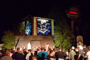 Eine nächtliche Außenansicht mit Menschen unten, Kindern auf der Bühne vor ihnen, einer braunen Wand hinter der Bühne, einem Gebäude mit Werbetafeln dahinter, Bäumen auf beiden Seiten des Gebäudes und Tafeln mit Text auf der rechten Seite.