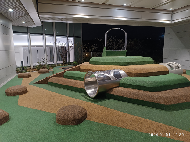 Ein Minigolf-Platz auf einem grünen Teppich in einem Raum mit Pflanzen, Deckenleuchten und Glaswänden im Hintergrund, mit einem Wasserzeichen unten.