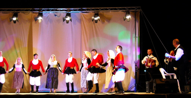 Performer auf der Bühne mit einem Vorhanghintergrund, Scheinwerfer oben, Musiker mit Instrumenten rechts und einem weißen Stuhl in der rechten unteren Ecke.