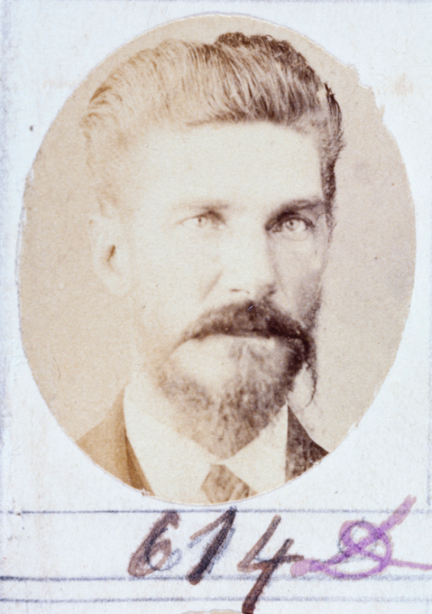 Ein altes Foto eines Mannes mit Bart und Schnurrbart, der direkt in die Kamera schaut und leicht geöffnete Lippen hat.