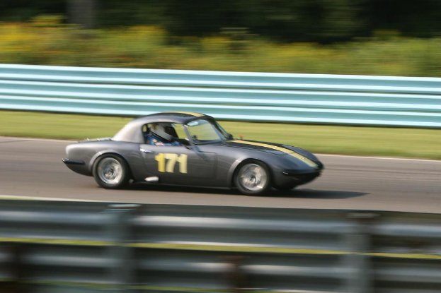 Ein Auto mit einem Helmträger fährt auf einer Straße entlang, die von Barrieren gesäumt ist, mit Bäumen im Hintergrund.