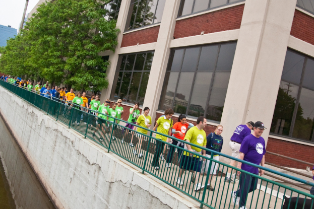 Ein paar Menschen spazieren auf der rechten Seite eines Weges entlang einer Reling, mit einem Gebäude mit Glasfenstern und Bäumen im Hintergrund und einem See auf der linken Seite unter einem klaren Himmel.