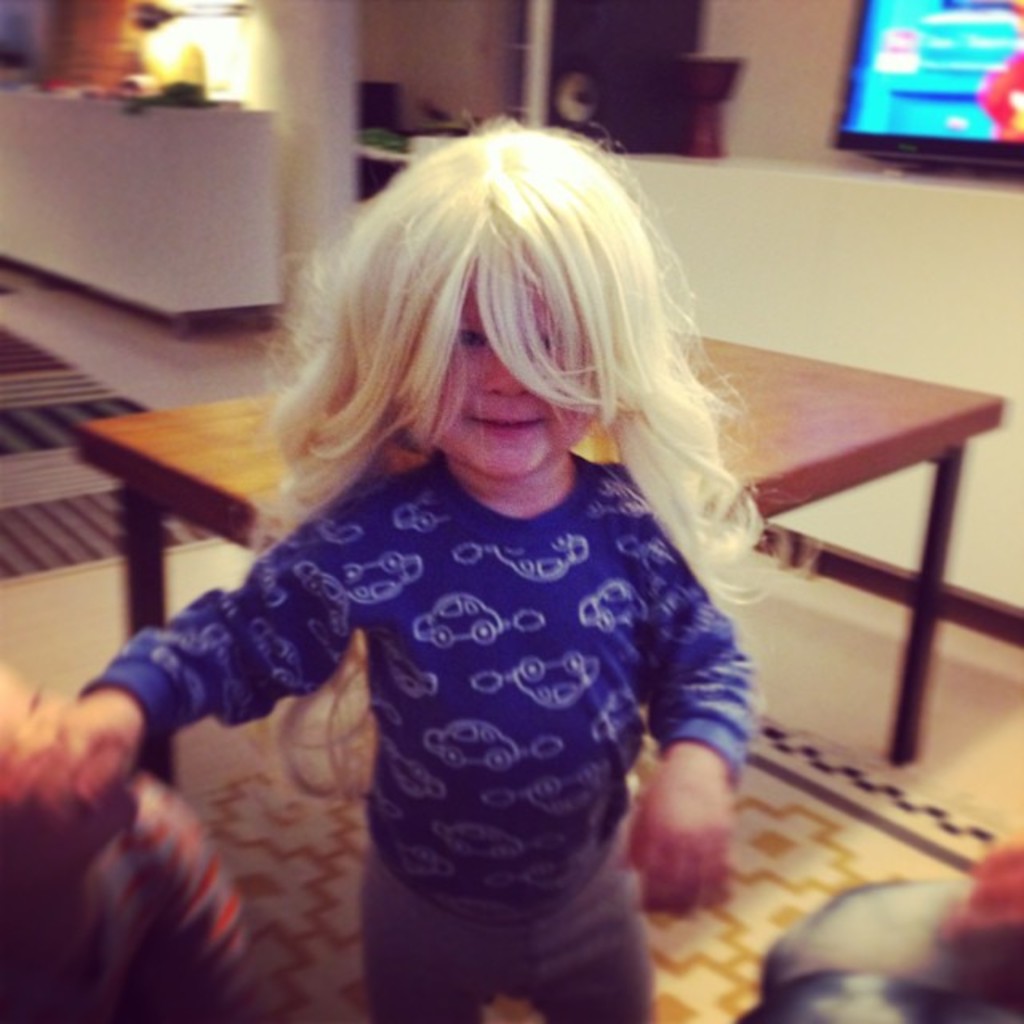 Ein kleines Mädchen mit langen weißen Haaren, das eine blaue T-Shirt und graue Hosen trägt, steht vor einem Tisch, mit einem weißen Schreibtisch, Fernseher und Lautsprecher im Hintergrund und einem braunen Teppich auf dem Boden.