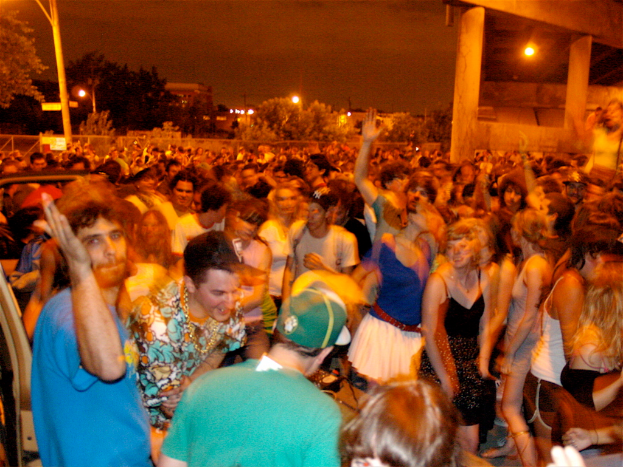 Menschen tanzen im Vordergrund auf einem Konzert, mit Lichtern, Pfählen, Bäumen und anderen Gegenständen im Hintergrund.