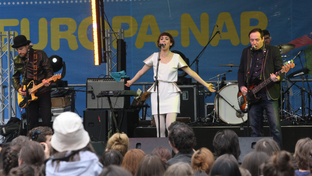Ein Konzertauftritt mit vier Personen auf der Bühne: eine Frau in einem weißen Kleid, die in der Mitte singt, zwei Männer, die links und rechts Gitarren spielen, und eine Menge vor einer Wand mit Lautsprechern im Hintergrund.