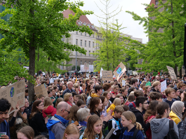 Eine große Menschenmenge protestiert vor einem Gebäude in Berlin, hält Schilder und steht in der Nähe von Bäumen, Fahrzeugen und einem Lautsprecher.