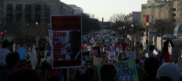 Menschen mit Plakaten auf einer Straße protestierend, mit Gebäuden, Bäumen, Laternen und Himmel im Hintergrund.