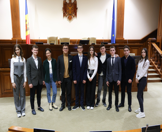 Gruppe strahlender Menschen vor Gerichtssaal mit Monitoren und Mikrofonen auf Tischen, vor einer hölzernen Plattform mit Flaggen, Stühlen und einer wandmontierten Einrichtung und Treppe mit Geländern auf der rechten Seite.