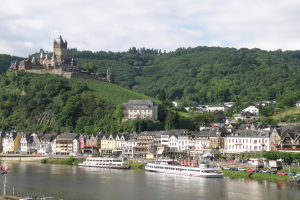 Ein malerischer Blick auf den Rhein in Deutschland, mit einer Burg auf einem Hügel im Hintergrund, Booten auf dem Fluss, Fahrzeugen auf einer nahen Straße und einem bewölkten Himmel.